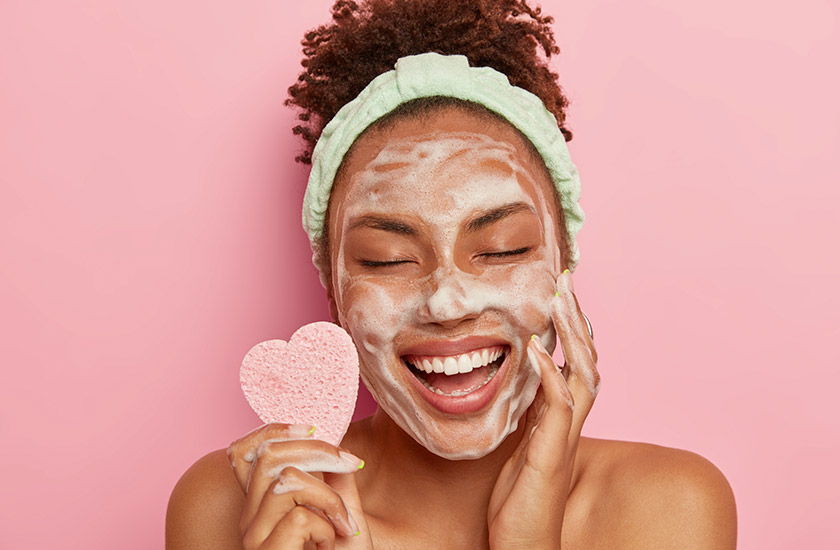mulher negra com uma expressão de felicidade no rosto, realizando todo passo a passo skincare e com uma esponja em forma de coração rosa na mão