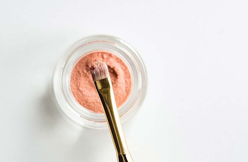 pincel de maquiagem pegando uma pequena quantidade de um recipiente com blush em pó rosa