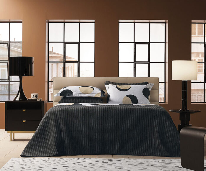 Cama de casal com lençol preto, dois travesseiros pretos, dois travesseiros com estampa de círculos por cima em um ambiente claro e janelas amplas