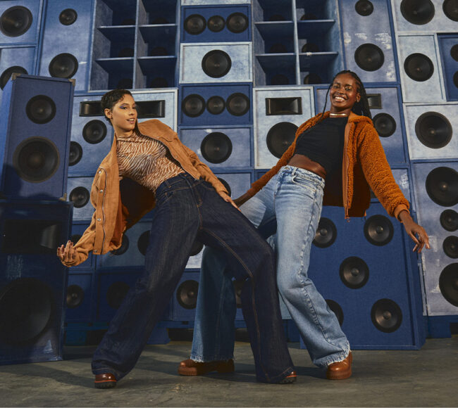 Duas mulheres em pose de dança vestindo calça jeans em um fundo azul com várias caixas de som 